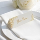 Personalised wedding place card with your name in elegant design on folded white cardstock. Positioned on a neatly folded white napkin atop a white plate, with a soft-focus white rose in the background.