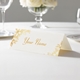 Personalised wedding place card with your name in elegant design on folded white cardstock. Displayed on a white tablecloth with wine glasses, plates, and a floral centrepiece in the softly blurred background.