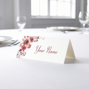 Personalised wedding place card with your name in elegant design on folded white cardstock. Placed on a table set for a formal event, with plates, cutlery, and wine glasses in the softly blurred background.