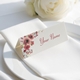 Personalised wedding place card with your name in elegant design on folded white cardstock. Positioned on a neatly folded white napkin atop a white plate, with a soft-focus white rose in the background.