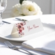 Personalised wedding place card with your name in elegant designt on folded white cardstock. Placed on a white plate with silver cutlery and a neatly folded napkin, with soft-focus white flowers in the background.