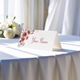 Personalised wedding place card with your name in elegant design on folded white cardstock. Displayed on a table with a flowing white tablecloth, with soft natural light and white roses in the background.