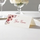 Personalised wedding place card with your name in elegant design on folded white cardstock. Displayed on a white tablecloth with wine glasses, plates, and a floral centrepiece in the softly blurred background.