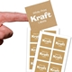 Close-up of a hand holding a square kraft label with white print reading White Print Kraft Labels above two sheets of matching stickers. The labels have a striped brown texture and are arranged neatly on white backing sheets, ideal for customised branding.