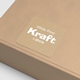 Square personalised kraft label with white print reading White Print Kraft Labels applied to a light brown cardboard box. The sticker features a striped texture and rounded corners, showing how the custom label looks when used on packaging.