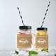 Two mason jar drinks with black striped straws and customised 60mm square kraft labels reading White Print Kraft Labels, 12 Stickers Per Sheet. One jar contains pink fruit and ice, the other lemon slices. Both labels have a brown striped texture and are set against a light background with scattered ice and mint.