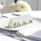 blank wedding place card with elegant design on folded white cardstock. Placed on a white plate with silver cutlery and a neatly folded napkin, with soft-focus white flowers in the background.