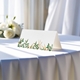 blank wedding place card printed with an elegant design on folded white cardstock ready to write your names on. Displayed on a table with a flowing white tablecloth, with soft natural light and white roses in the background.