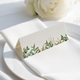 wedding place card with an elegant design on folded white cardstock, ready to write your names on. Positioned on a neatly folded white napkin atop a white plate, with a soft-focus white rose in the background.