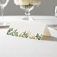 wedding place card with an elegant design on folded white cardstock ready to write your name on. Displayed on a white tablecloth with wine glasses, plates, and a floral centrepiece in the softly blurred background.