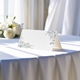 blank wedding place card printed with an elegantdesign on folded white cardstock ready to write your names on. Displayed on a table with a flowing white tablecloth, with soft natural light and white roses in the background.