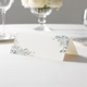 wedding place card with an elegant design on folded white cardstock ready to write your name on. Displayed on a white tablecloth with wine glasses, plates, and a floral centrepiece in the softly blurred background.