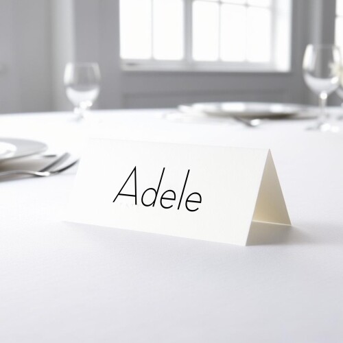 Personalised wedding place card with your name in elegant black script on folded white cardstock. Placed on a table set for a formal event, with plates, cutlery, and wine glasses in the softly blurred background.