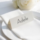 Personalised wedding place card with your name in elegant black script on folded white cardstock. Positioned on a neatly folded white napkin atop a white plate, with a soft-focus white rose in the background.