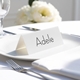 Personalised wedding place card with your name in elegant black script on folded white cardstock. Placed on a white plate with silver cutlery and a neatly folded napkin, with soft-focus white flowers in the background.