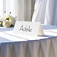 Personalised wedding place card with your name in elegant black script on folded white cardstock. Displayed on a table with a flowing white tablecloth, with soft natural light and white roses in the background.