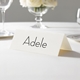 Personalised wedding place card with your name in elegant black script on folded white cardstock. Displayed on a white tablecloth with wine glasses, plates, and a floral centrepiece in the softly blurred background.