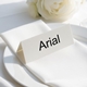 Personalised wedding place card with your name in elegant black script on folded white cardstock. Positioned on a neatly folded white napkin atop a white plate, with a soft-focus white rose in the background.