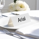 Personalised wedding place card with your name in elegant black script on folded white cardstock. Placed on a white plate with silver cutlery and a neatly folded napkin, with soft-focus white flowers in the background.