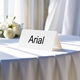 Personalised wedding place card with your name in elegant black script on folded white cardstock. Displayed on a table with a flowing white tablecloth, with soft natural light and white roses in the background.