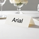 Personalised wedding place card with your name in elegant black script on folded white cardstock. Displayed on a white tablecloth with wine glasses, plates, and a floral centrepiece in the softly blurred background.