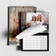 Two wall calendars displayed, one showing a woman posing in front of festive lights, and the other partially open to a month view with a photo of two smiling women in white tops and sunglasses, standing outdoors. Both calendars are wire-bound with black borders and a central hanging hook, set against a light background.