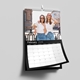 February 2026 wall calendar page featuring a photo of two smiling women in white tops and jeans standing outdoors in a city setting. The calendar has a black header with white text and a clean grid layout below the image. Wire-bound with a central hanging hook, the calendar is shown partially open against a light grey wall.