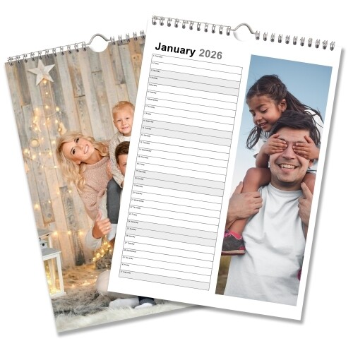 A 2026 personalised wall planner calendar is shown open to January. The layout includes a vertical list of dates with space for notes beside each day and a full-length photo of a girl playfully covering a smiling man's eyes. Behind it, the cover features a family posing in front of a festive wooden backdrop with fairy lights. The calendar is spiral-bound with a central hanging hook.