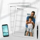 Two 2026 wall planner calendars are displayed on a white surface. The March page features a smiling father hugging his young son beside a scenic waterfront. Behind it, the April page is partially visible, showing a lined daily layout. A smartphone and black pen are placed nearby, adding a realistic desk setup. Both calendars are wire-bound with a hanging hook at the top.