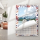Wall calendar with a colourful leaf and coral-themed border hanging on a white wall in a modern room. The calendar features a scenic photo of two people standing by a lake with snow-capped mountains in the background. A partially turned page reveals a monthly grid, suggesting it is transitioning between months.
