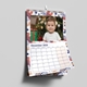 Wall calendar open to December 2026, featuring a young child in front of a decorated Christmas tree with presents. The page has a festive abstract border in red, blue, and beige, and includes a clear monthly grid with bold dates and weekdays. The calendar hangs neatly on a white wall using a metal hook.