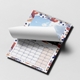 Open wall calendar with a vibrant red, blue, and beige abstract border. The current page is being turned, partially revealing a scenic sky photo above and the full calendar grid for May 2026 below. The calendar is wire-bound with a central hanging hook and rests on a light surface.