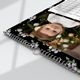 Close-up of a floral-themed wall calendar showing a smiling baby in front of a decorated Christmas tree. The calendar is spiral-bound with a silver metal hanging hook, and the edge of the date grid is visible in the corner. The design features a black background with purple and white flowers and green leaves.