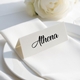 Personalised wedding place card with your name in elegant black script on folded white cardstock. Positioned on a neatly folded white napkin atop a white plate, with a soft-focus white rose in the background.