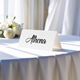 Personalised wedding place card with your name in elegant black script on folded white cardstock. Displayed on a table with a flowing white tablecloth, with soft natural light and white roses in the background.