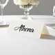 Personalised wedding place card with your name in elegant black script on folded white cardstock. Displayed on a white tablecloth with wine glasses, plates, and a floral centrepiece in the softly blurred background.