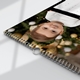 Close-up of an A3 wall calendar with a sleek black design, showing a photo of a young child in front of a Christmas tree. The calendar has spiral binding with a metal hanging hook and part of a monthly grid visible.