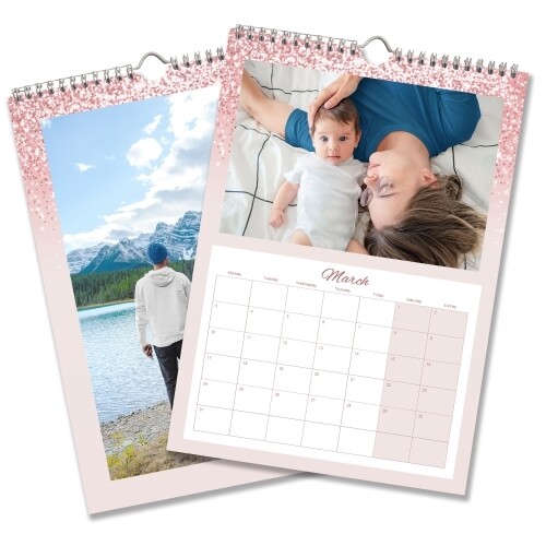 Two A4 wall calendars with pink glitter borders. One features a landscape photo of a person by a lake and mountains, and the other shows March 2025 with a photo of a parent and baby lying on a bed. Both calendars have monthly grids, spiral bindings, and hanging hooks.