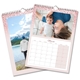 Two A4 wall calendars with pink glitter borders. One features a landscape photo of a person by a lake and mountains, and the other shows March 2025 with a photo of a parent and baby lying on a bed. Both calendars have monthly grids, spiral bindings, and hanging hooks.