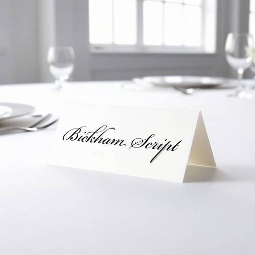 Personalised wedding place card with your name in elegant black script on folded white cardstock. Placed on a table set for a formal event, with plates, cutlery, and wine glasses in the softly blurred background.