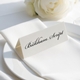 Personalised wedding place card with your name in elegant black script on folded white cardstock. Positioned on a neatly folded white napkin atop a white plate, with a soft-focus white rose in the background.