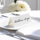 Personalised wedding place card with your name in elegant black script on folded white cardstock. Placed on a white plate with silver cutlery and a neatly folded napkin, with soft-focus white flowers in the background.