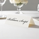 Personalised wedding place card with your name in elegant black script on folded white cardstock. Displayed on a white tablecloth with wine glasses, plates, and a floral centrepiece in the softly blurred background.