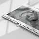 Close-up of an A4 wall calendar with a marble design, featuring a black and white photo of baby feet cradled by adult hands. The calendar has spiral binding, a metal hanging hook, and part of a monthly grid visible.