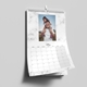 An A4 wall calendar for May 2025, featuring a marble design. The page displays a photo of a man carrying a child on his shoulders, with a monthly grid below. The calendar has spiral binding and a hanging hook, mounted on a grey wall.