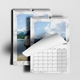 Two 2026 wall calendars with white marble borders are shown hanging side by side. One calendar is open to January with the page being turned, revealing a scenic sky and mountain image. The calendar grid for January is visible with a clean layout. The second calendar behind features a photo of a man in a white hoodie by a mountain lake. Both calendars are spiral-bound with central hanging hooks.
