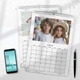 Two 2026 wall calendars with white marble borders lie flat on a white surface. The top calendar is open to April, featuring a photo of two children in white shirts—one wearing a straw hat—posed in front of pastel balloons. The calendar grid displays all days of April. The second calendar beneath shows part of a photo of a child rollerblading at the beach. A smartphone and pen rest nearby, creating a tidy workspace setting.