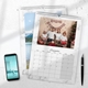 n A3 wall calendar with a marble design, open to December 2025. The page features a family Christmas photo with festive decorations. A second calendar showing a lake and mountain scene is partially visible underneath. A smartphone and pen are placed beside the calendars.