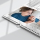 A close-up of a 2026 wall calendar with a white marble border, lying flat on a white surface. The featured photo shows a parent in a blue shirt lying on a bed, gently cradling their baby. The spiral binding and central hanging hook are clearly visible, highlighting the calendar's practical design. The corner of the calendar grid is partially visible, showing part of the June layout.
