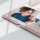 Close-up of an A3 calendar with a pink glitter border. The visible page features a photo of three people in red capes and masks, smiling. The calendar is wire-bound with a hanging loop, lying flat on a white surface.